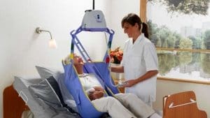 Nurse uses Arjo Maxi Sky 440 Portable Ceiling Lift with blue sling for elderly patient mobility support.